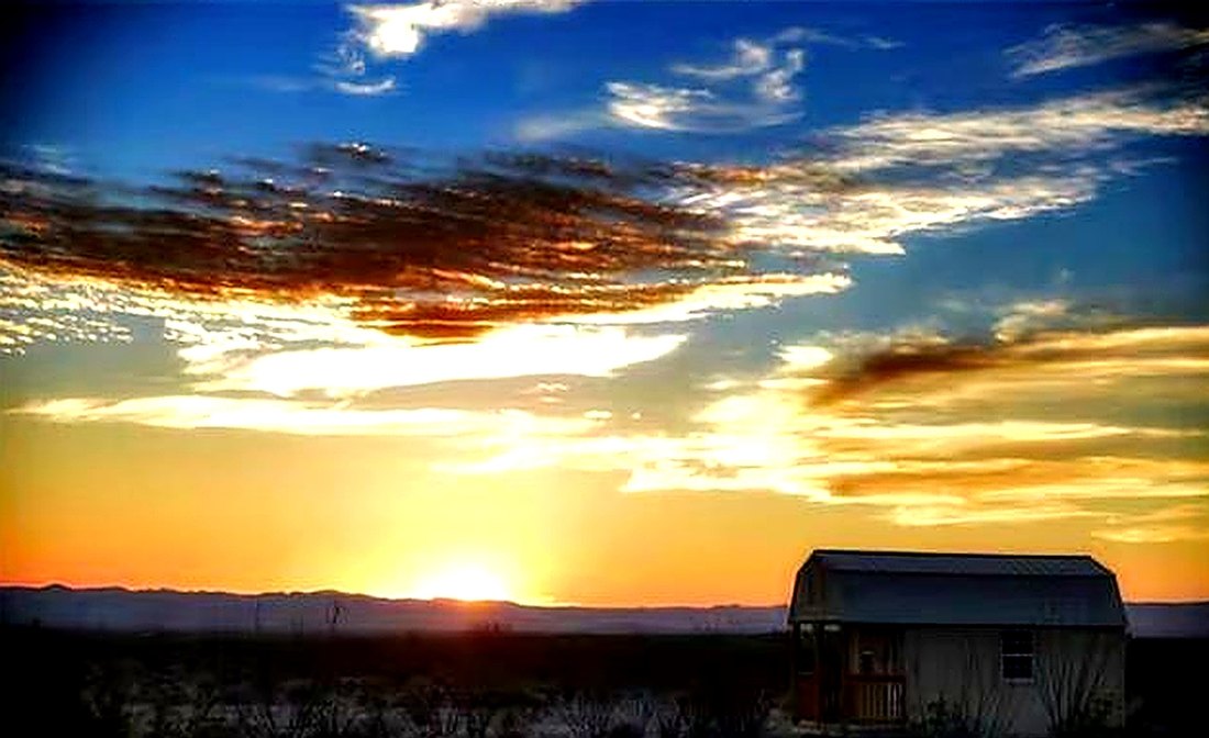 A wide desert sunset with layered orange clouds above the silhouetted cabin